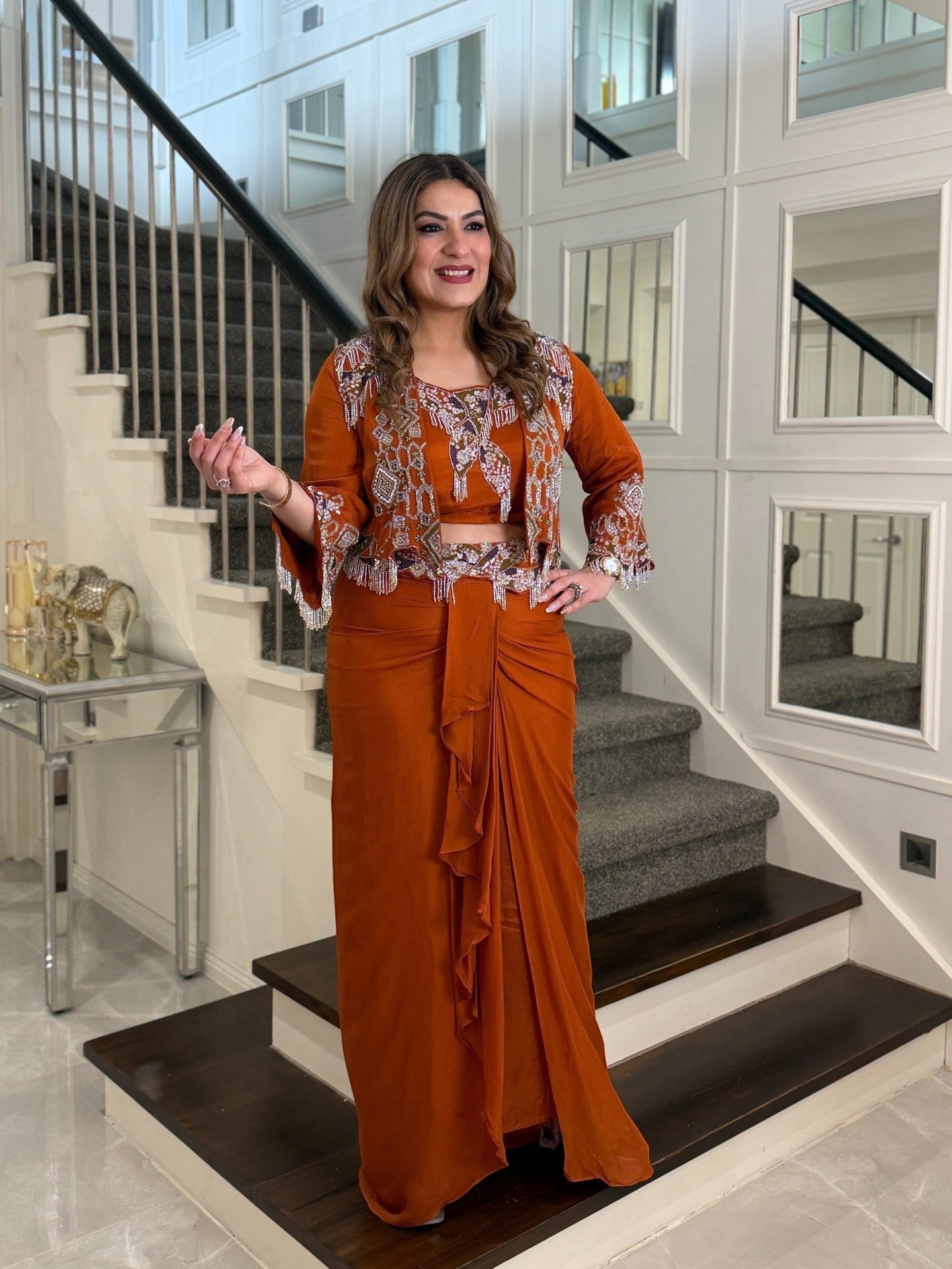 Woman wearing a rust-orange PanacheByDivya indo-western outfit with an embroidered jacket and draped skirt.